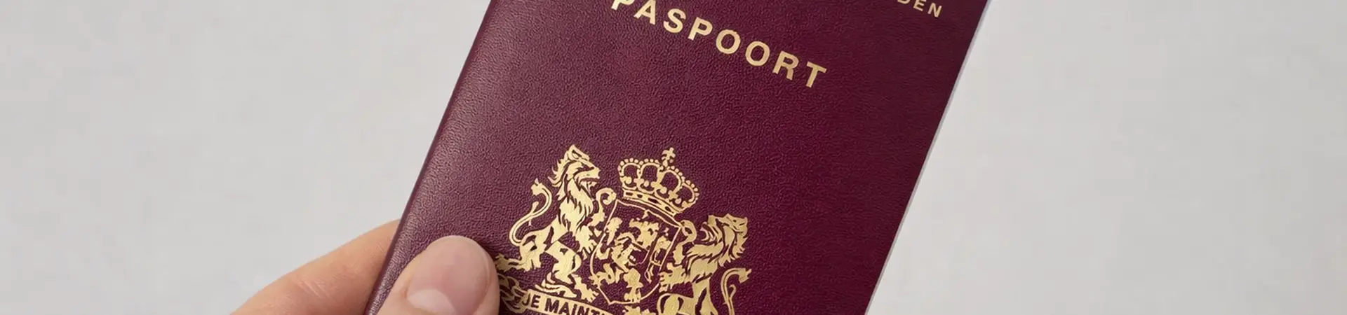 A hand holding a Netherlands passport with a maroon cover, featuring a gold emblem and text in Dutch.