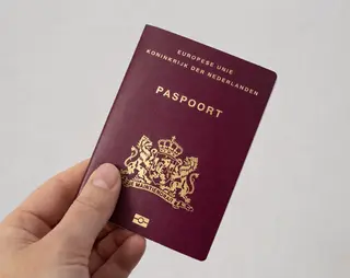 A hand holding a Netherlands passport with a maroon cover, featuring a gold emblem and text in Dutch.