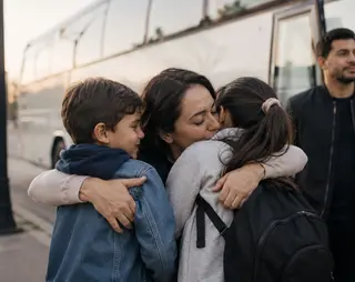 A woman hugs two children near a bus, with a man observing in the background.