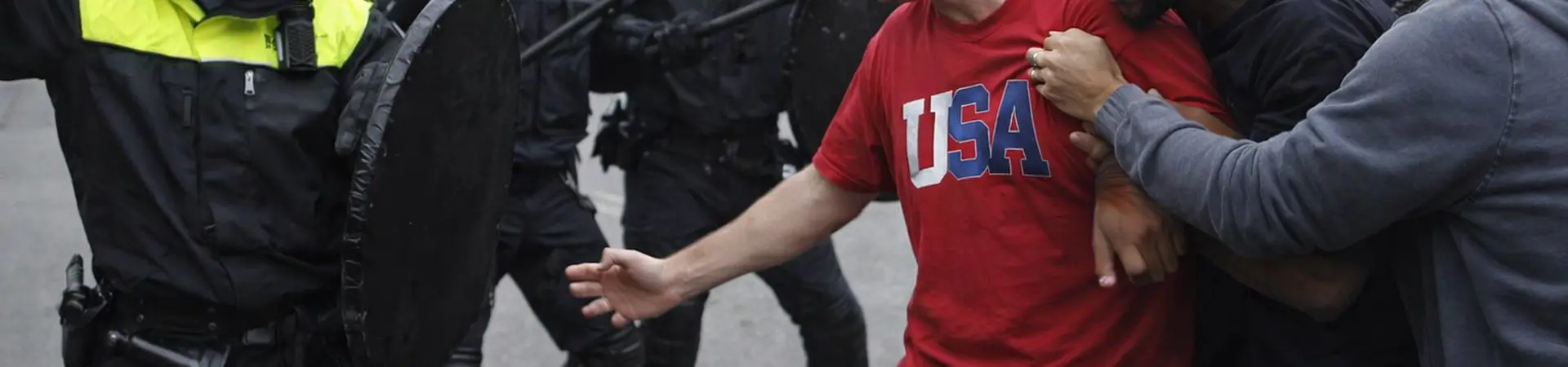 Protesters confront police in riot gear on a city street. One man, wearing a "USA" shirt, is held back by others, amid a tense standoff.