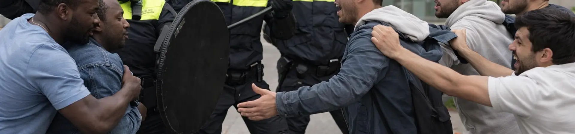 Protest scene with police in riot gear confronting a group of men. Police are holding shields and batons, while the men shout and gesture.