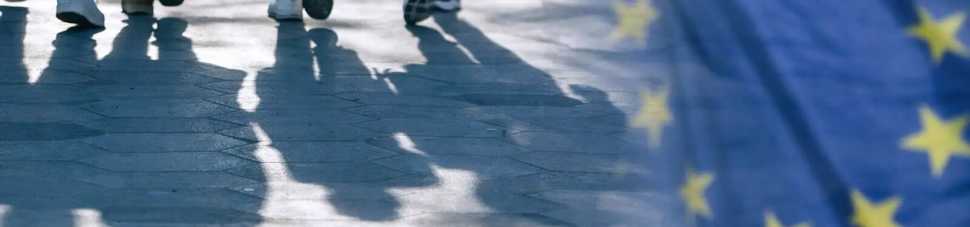 People walking on a sunlit pavement, casting shadows. The European Union flag is prominent on the right side of the image.
