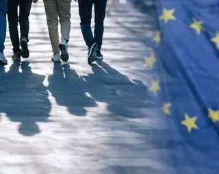 People walking on a sunlit pavement, casting shadows. The European Union flag is prominent on the right side of the image.