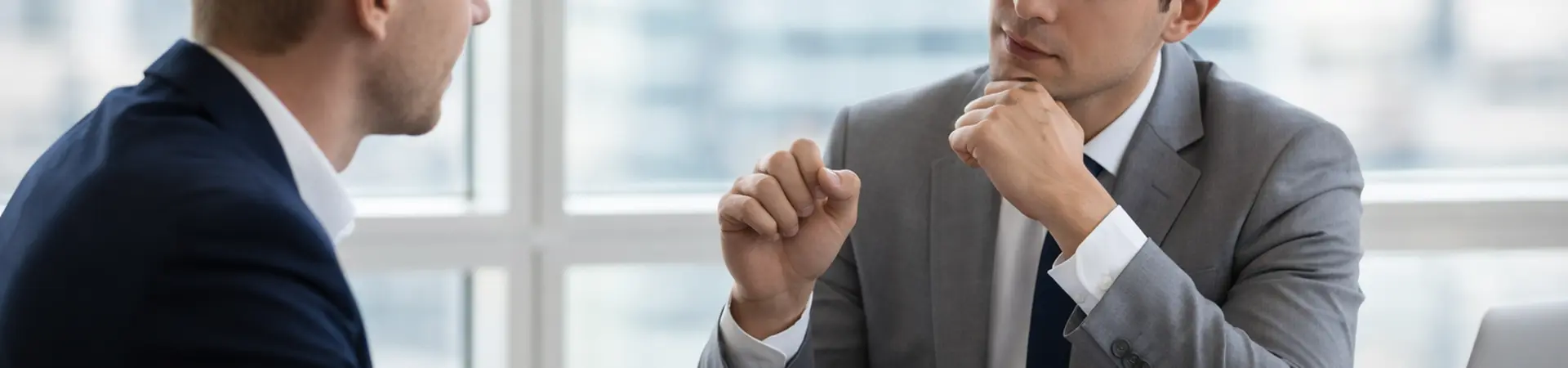 Two men in business attire engaged in a serious discussion at a table with documents, a laptop, and a cup of coffee in a bright office setting.