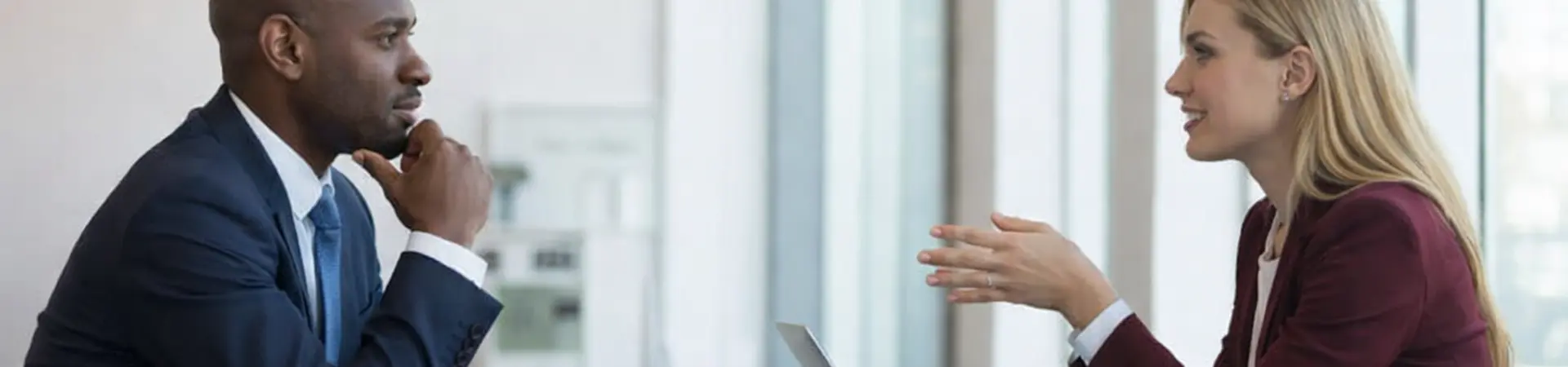 A man and a woman in business attire have a discussion across a table with a laptop, in a modern office setting with large windows.