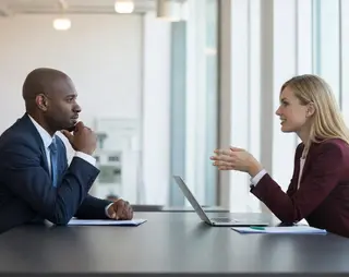 A man and a woman in business attire have a discussion across a table with a laptop, in a modern office setting with large windows.