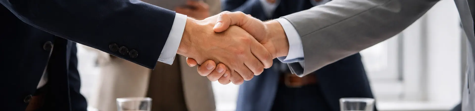 Two people in business attire shaking hands over a table with a contract, surrounded by colleagues applauding.