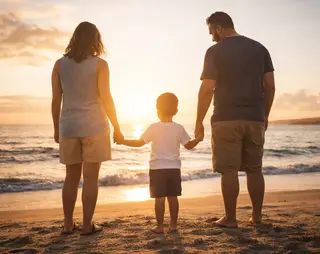 A family of three holds hands, standing on a beach at sunset, facing the ocean.