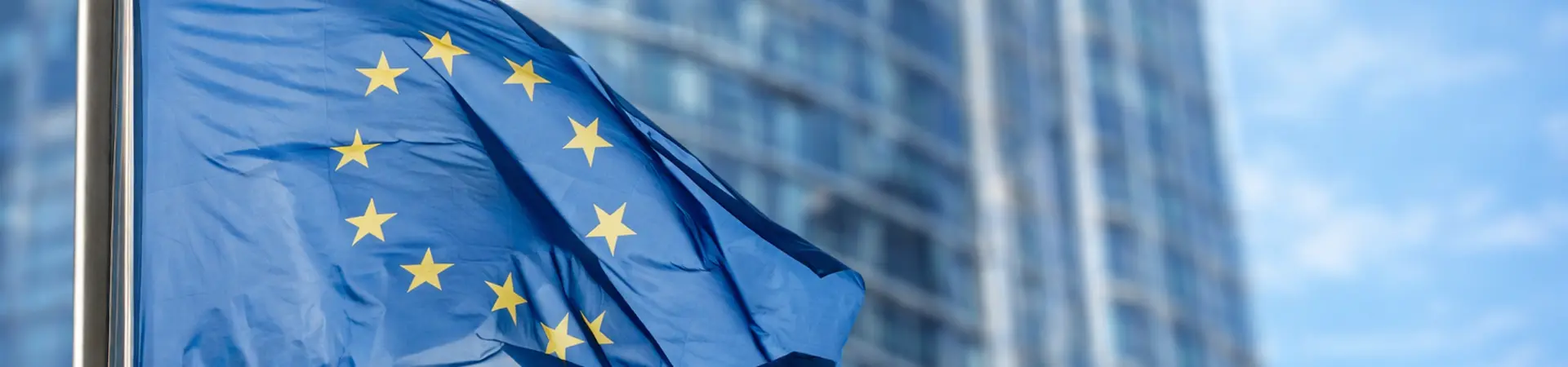 European Union flag waving in front of a modern glass building under a clear blue sky.