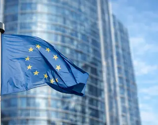 European Union flag waving in front of a modern glass building under a clear blue sky.