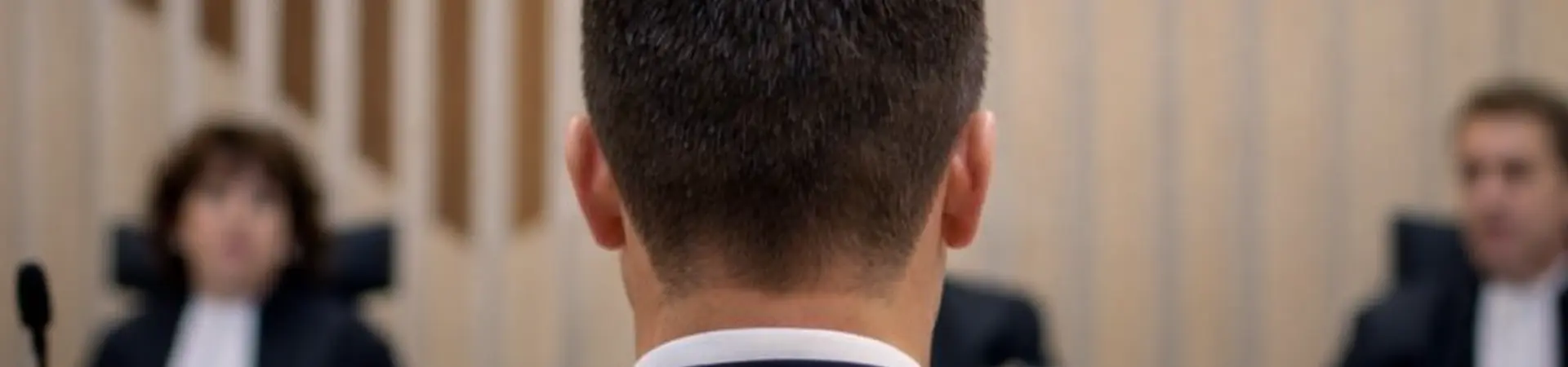 Rear view of a man in a suit facing three judges in a courtroom, with a wooden backdrop and a bench.
