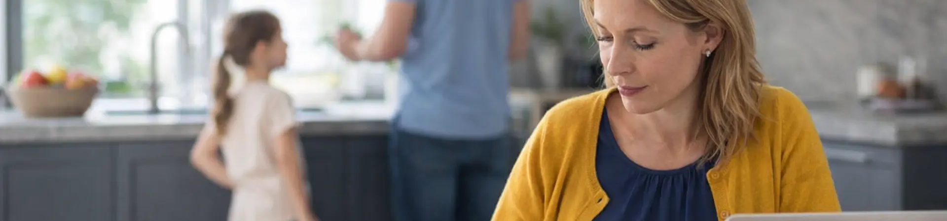 Woman working at kitchen table with a laptop and papers, while a man and child stand in the background near the kitchen counter.