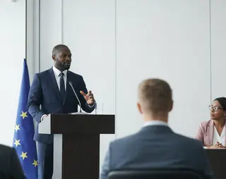 Man in a suit speaking at a podium with an EU flag, addressing a seated audience in a conference room.
