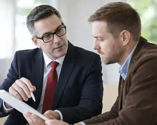 Two men sitting at a table, discussing over a tablet. One is in a suit with glasses, and the other in a brown jacket, listening attentively.