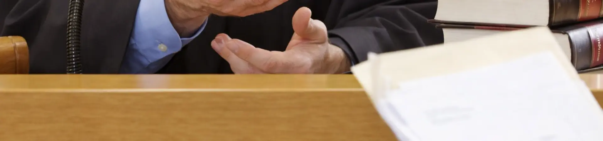 Judge gesturing with hands in courtroom, sitting at a bench with legal books, while a person holds documents in the foreground.