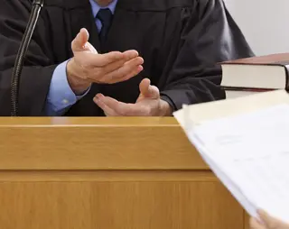 Judge gesturing with hands in courtroom, sitting at a bench with legal books, while a person holds documents in the foreground.
