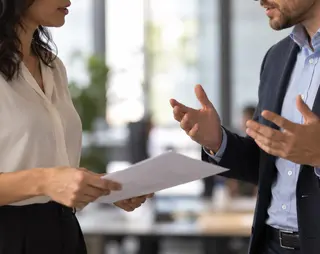 Two professionals engaged in a discussion, holding documents, standing in an office setting.