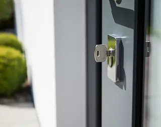 Een sleutel die in het sleutelgat van een grijze voordeur zit met struiken op de achtergrond