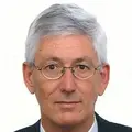 Een man van middelbare leeftijd met grijs haar en een bril, gekleed in een pak met een wit overhemd en een blauwe stropdas, tegen een effen witte achtergrond.