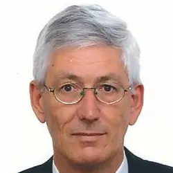 Een man van middelbare leeftijd met grijs haar en een bril, gekleed in een pak met een wit overhemd en een blauwe stropdas, tegen een effen witte achtergrond.