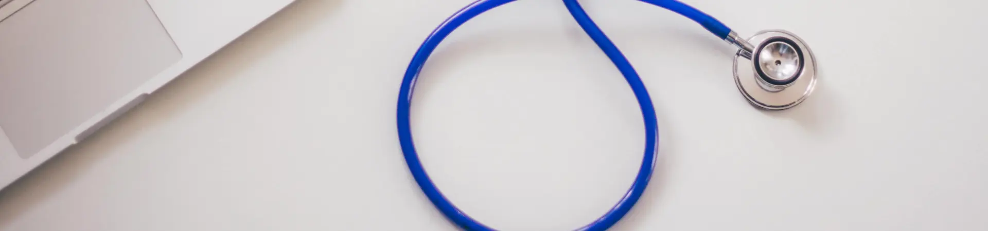 Op een wit bureau ligt een laptop en daarnaast een blauwe stethoscoop. 