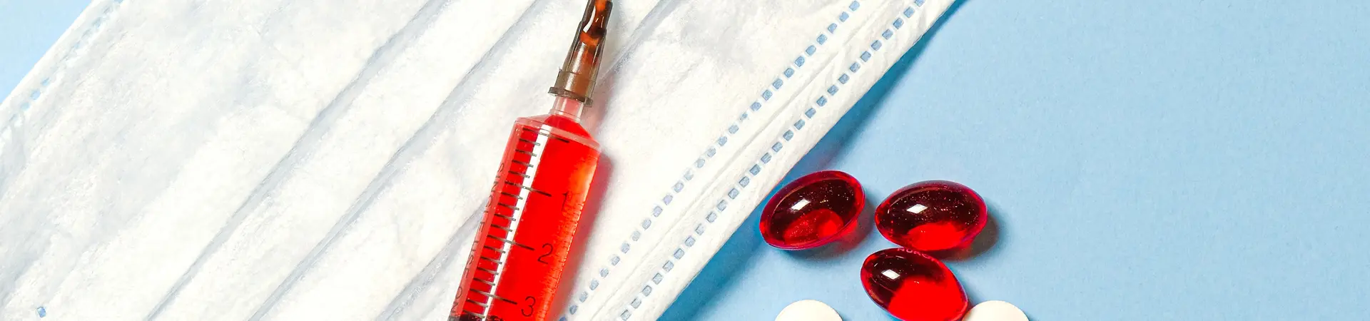 injectiespuit met rode vloeistof, rode cpsules en witte tabletten op een wit mondkapje.
