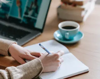 Iemand die in een notitieboekje schrijft naast haar een laptop en kop koffie