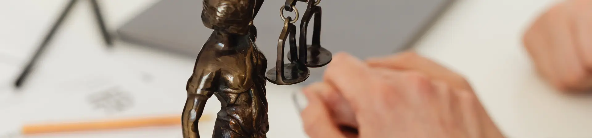 Vrouwe Justitia staat met haar weegschaal en zwaard op een bureau. Man en vrouw houden elkaars hand vast en zitten aan bureau.