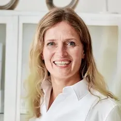 Smiling person with long blonde hair wearing a white shirt, standing indoors with a blurred background of shelves.