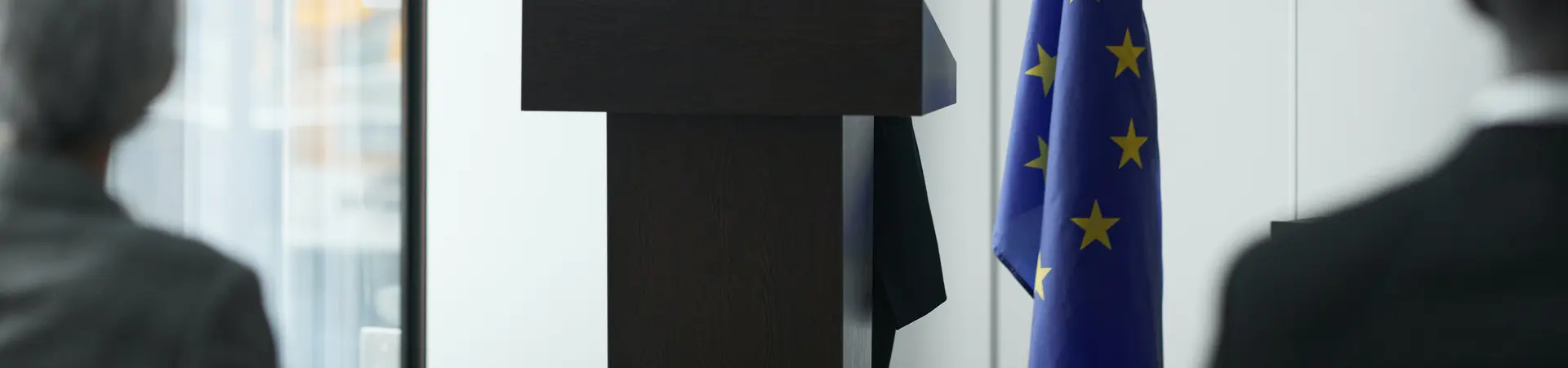 Een vrouw houdt een toespraak achter een desk. Achter de vrouw staat de vlag van de Europese Unie. Er zijn twee toeschouwers.