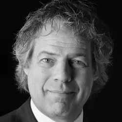 Man met krullend haar in een pak poseert voor een zwarte achtergrond, kijkt naar de camera.