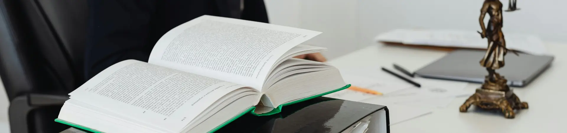 Man die achter zijn bureau zit met een boek, ordner, krant en tablet en een beeld van vrouwe Justitia