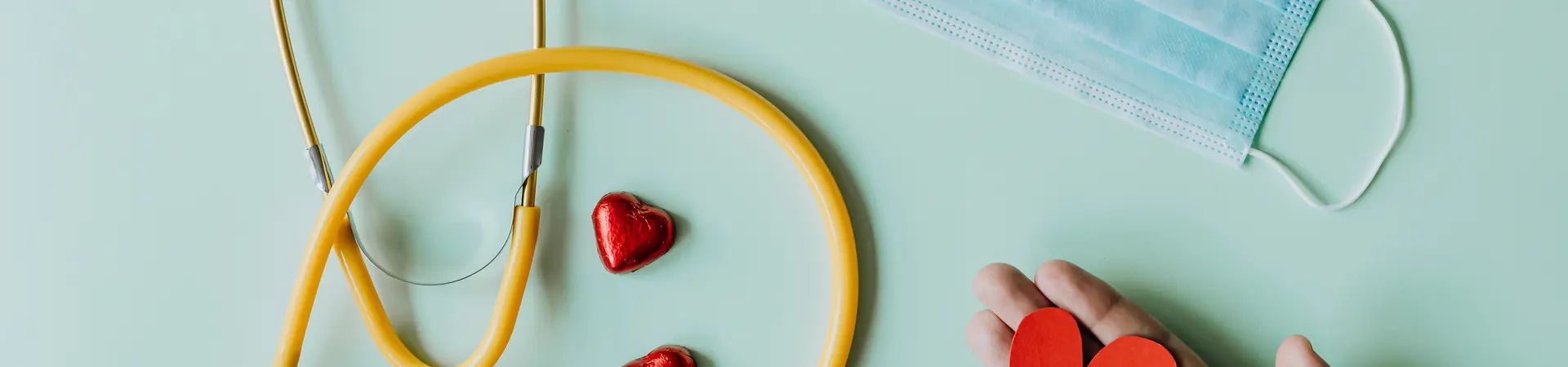 Gele stethoscoop, rode hartjes, een mondkapje en een hand met een papieren rood hartje