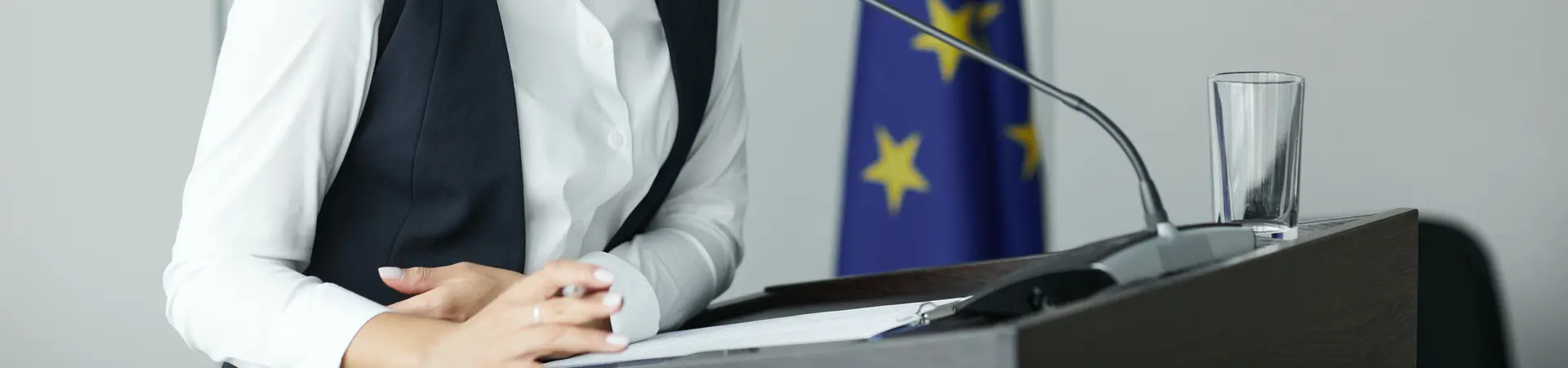 Een vrouw houdt een toespraak achter een desk. Achter de vrouw staat de vlag van de Europese Unie. 