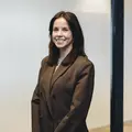 Vrouw met lang donker haar en een bruine blazer glimlacht in een kantooromgeving.