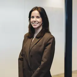 Vrouw met lang donker haar en een bruine blazer glimlacht in een kantooromgeving.
