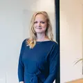 Vrouw met lang blond haar in een blauwe jurk staat glimlachend in een kantooromgeving.