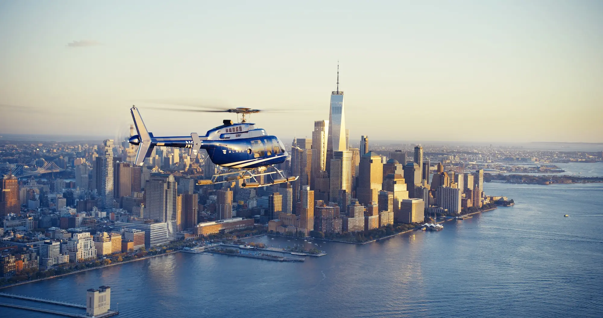 A blue helicopter flies over a cityscape with skyscrapers, including a tall tower, near a body of water during sunset.