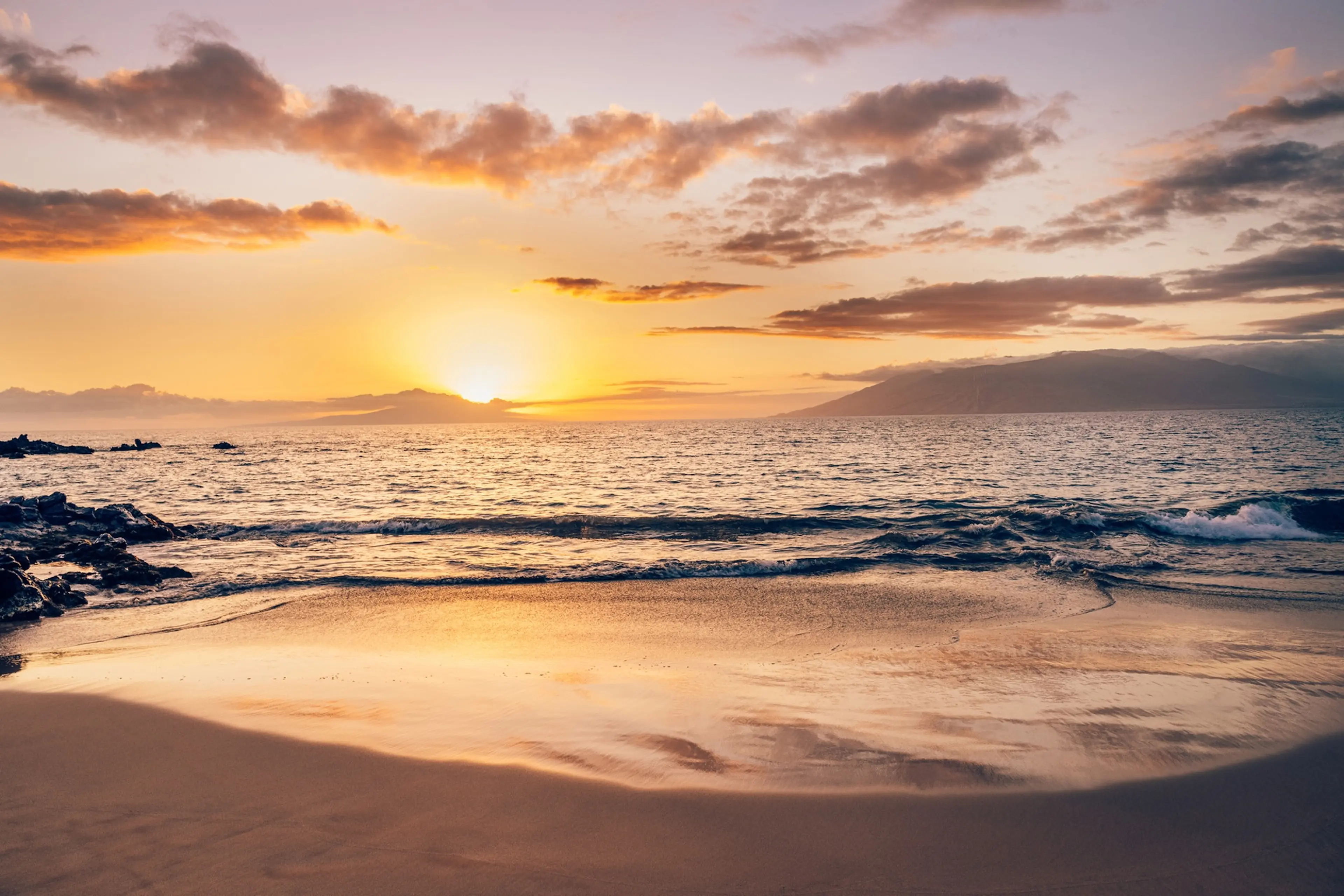 Sunset over a tranquil beach with gentle waves, golden reflections on wet sand, and a cloudy sky casting warm hues across the horizon.