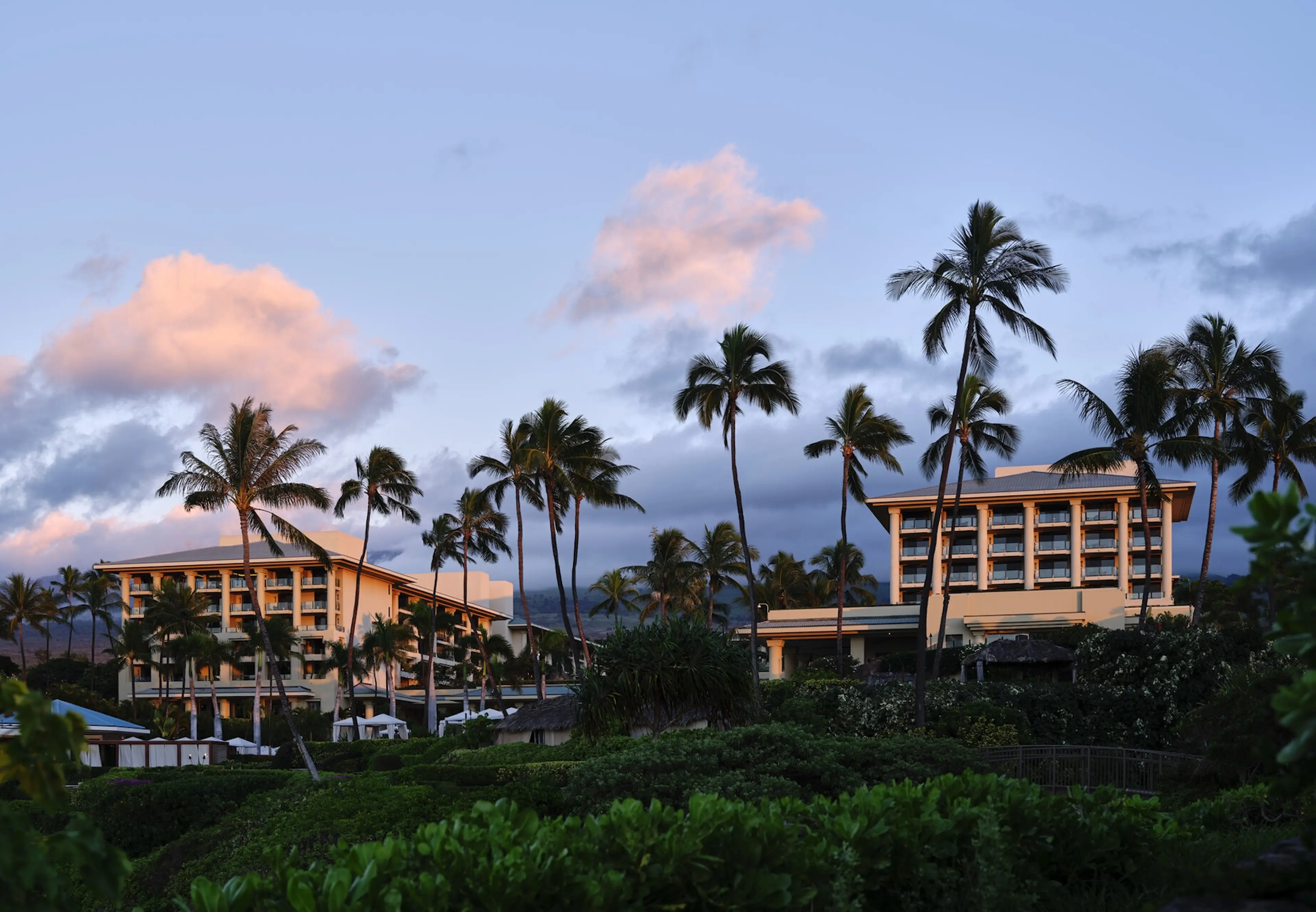Tropical resort buildings surrounded by palm trees at sunset, with pink clouds and a lush green foreground.