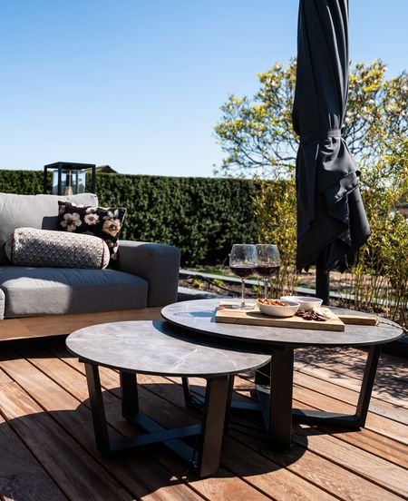 Outdoor patio with two tables, wine glasses, and snacks. Gray sofa with cushions. Rolled-up umbrella and greenery in the background.