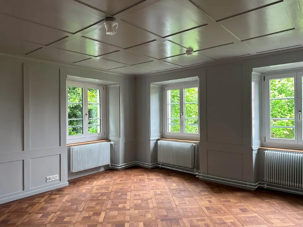 Ein Raum im Altbau mit zwei großen Fenstern, Parkettboden, perlgrauen Wänden und Deckenkasetten. Unter jedem Fenster befinden sich lackierte Heizkörper.