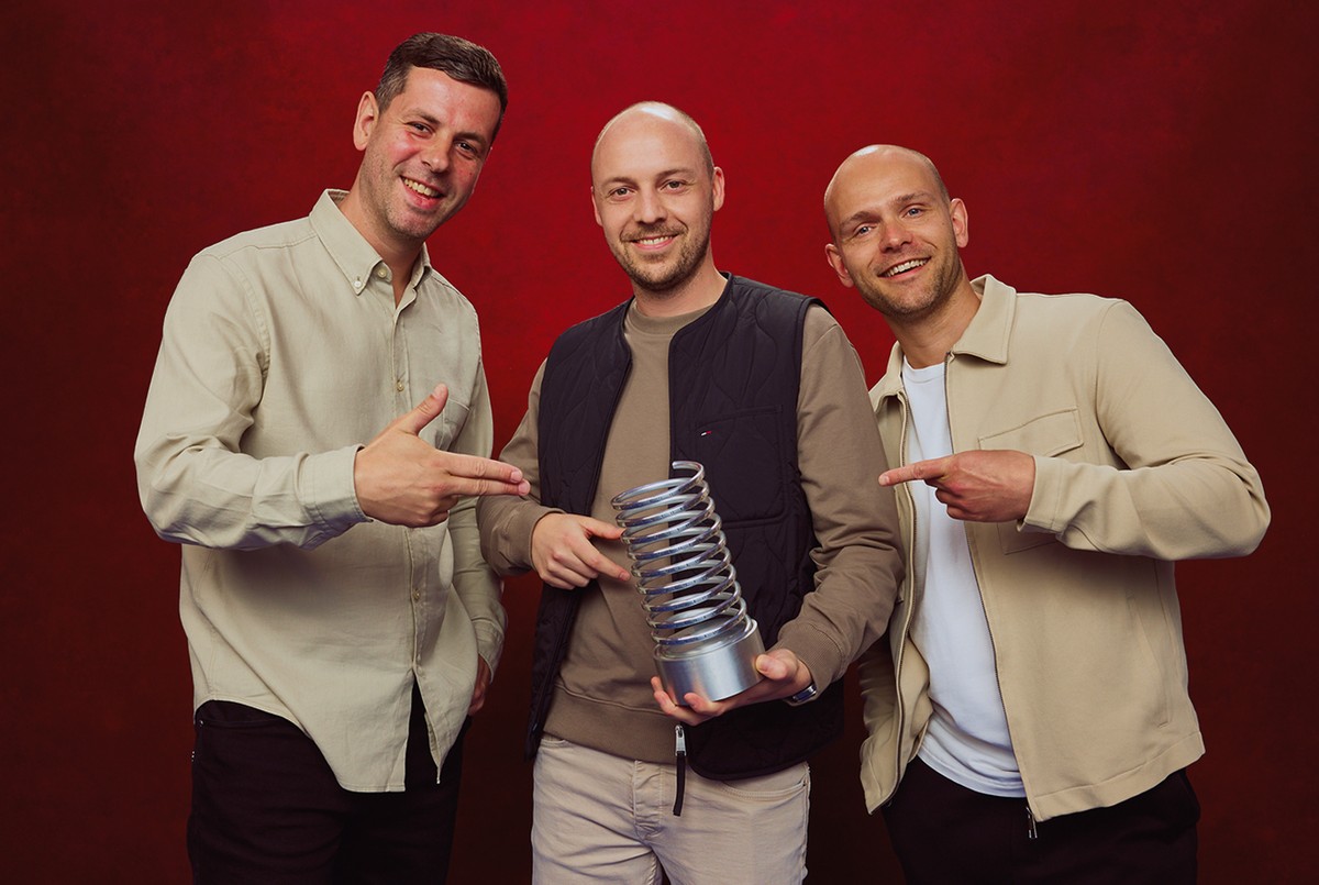 Three smiling men pose with a spiral trophy against a red backdrop, all wearing casual beige outfits.