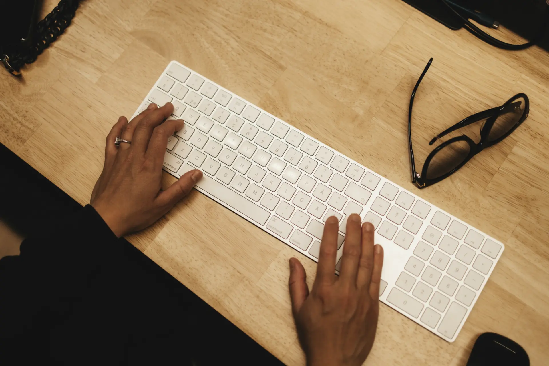 Hands typing on a white keyboard on a wooden desk, with glasses placed nearby.