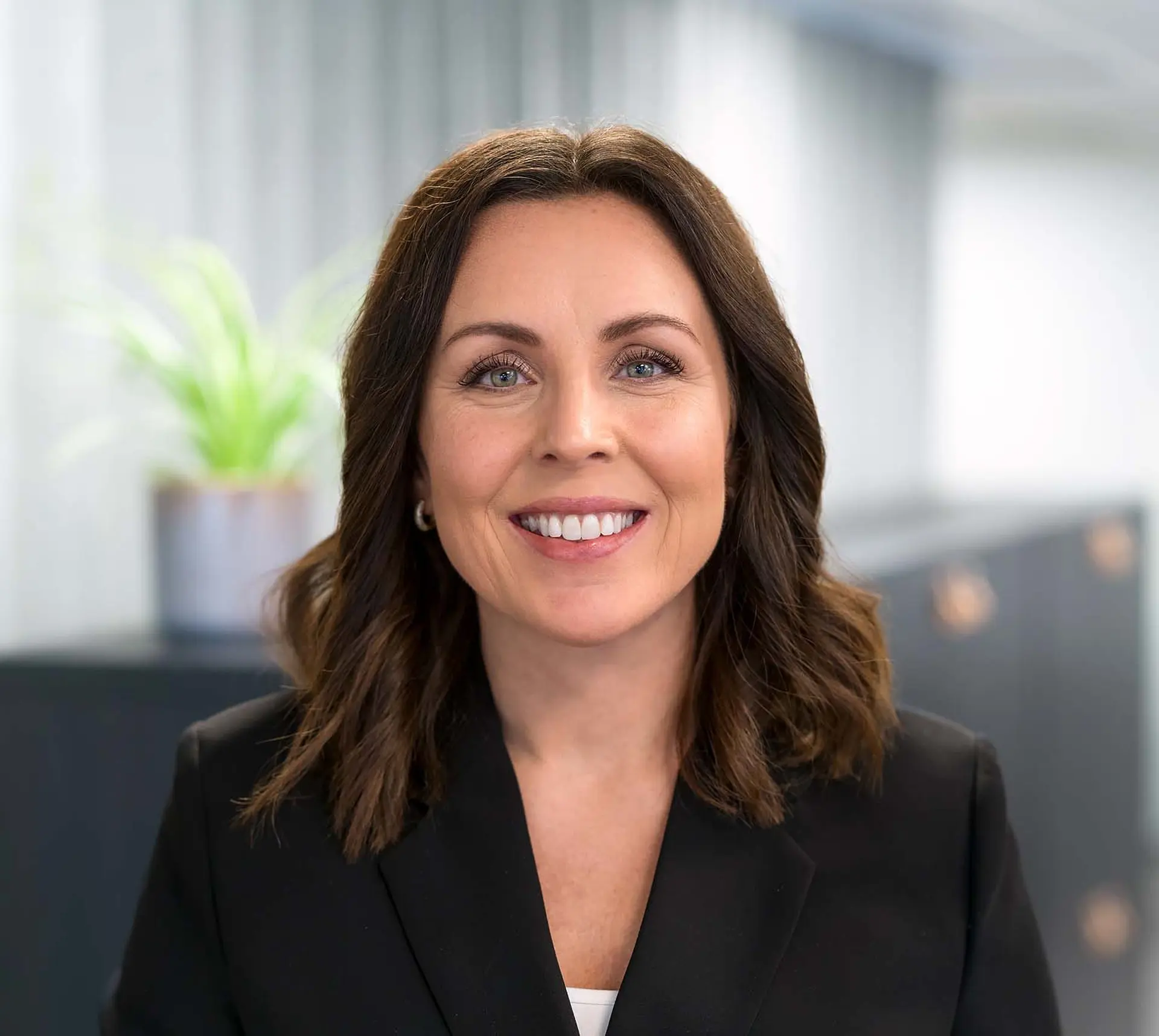 Smiling woman with brown hair in a black blazer, standing indoors with a blurred plant in the background.