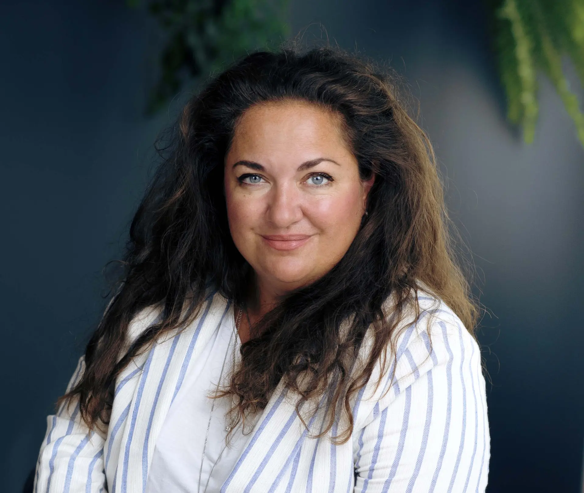 Woman with long dark hair in a striped blazer, smiling softly, against a dark backdrop with green foliage.