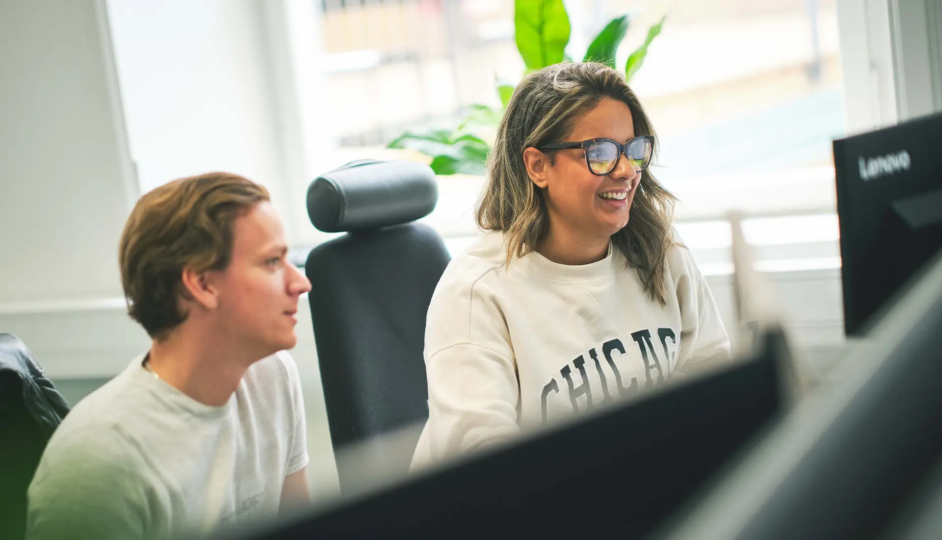 Two people working together at a desk with computers, smiling and looking at a screen, with plants in the background.