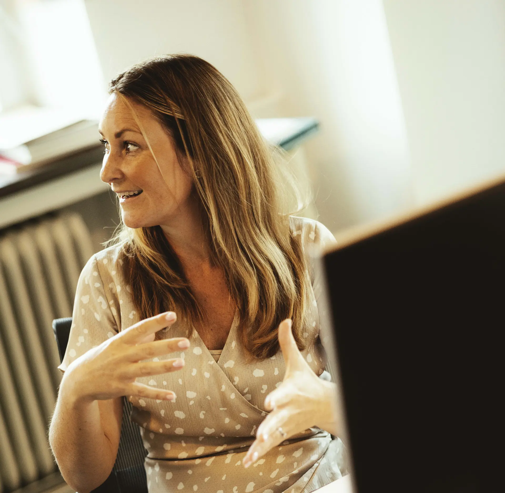 Woman with long hair gesturing while talking, seated indoors near a window with soft lighting and part of a computer screen visible in the foreground.