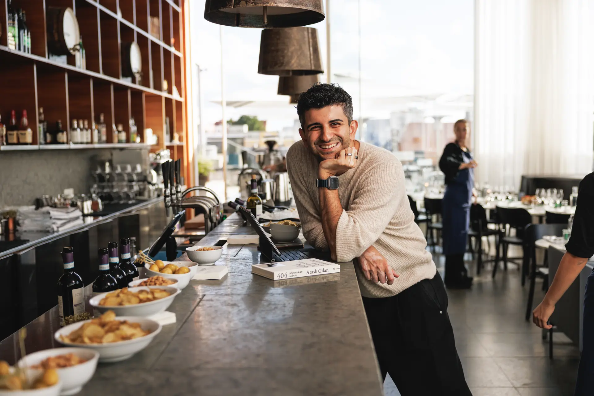 A man smiling and leaning on the counter in a restaurant bar, with bowls of snacks and wine bottles lined up, and a blurred person in the background.