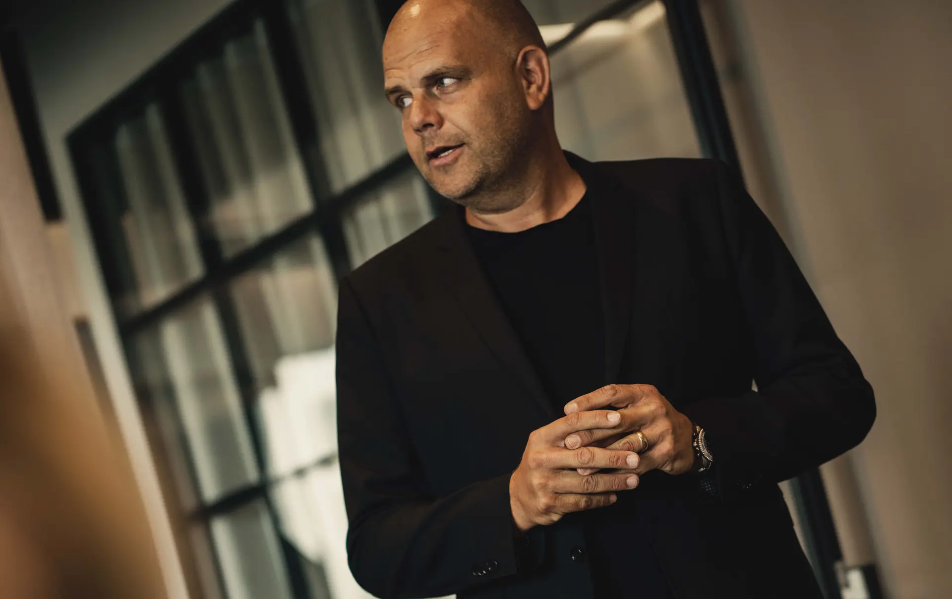 A bald man in a black suit stands in a modern office, looking to the side with hands held together.
